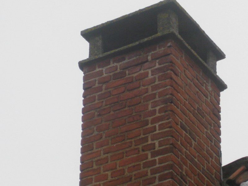 Nærbillede af en muret skorstenspibe i rød tegl med betonafdækning mod grå himmel.