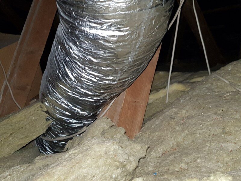 Nærbillede af en fleksibel ventilationsslange i sølvfarvet aluminium, der går gennem isolering på et tagrum.