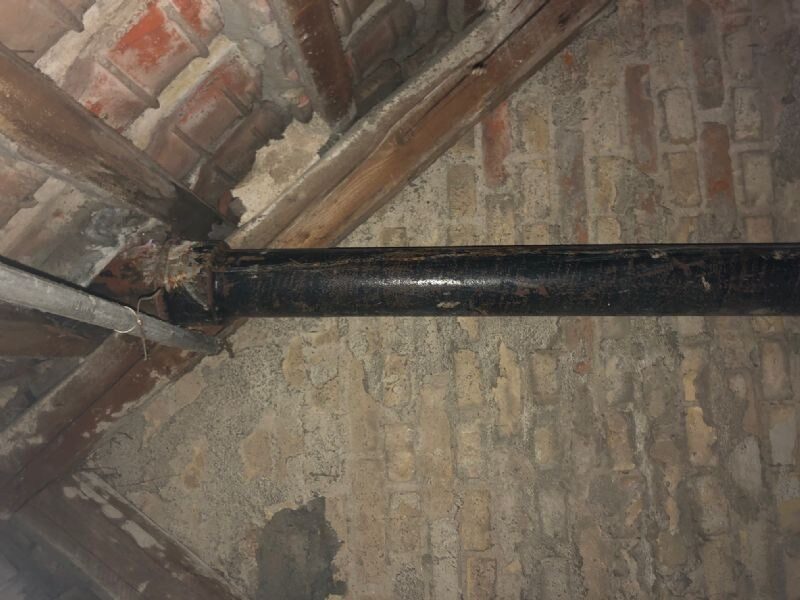 Nærbillede af en sort støbejernsrørgennemføring i et murstensgratspær, formentlig en gammel skorstens- eller ventilationsføring set indefra i tagrummet.