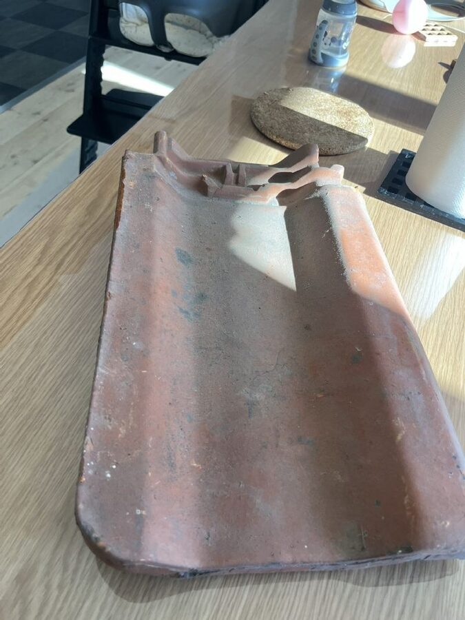 Enkelt tegltagsten fotograferet indendørs på et køkkenbord, med synlig ophængningsnæse og krakeleret overflade.