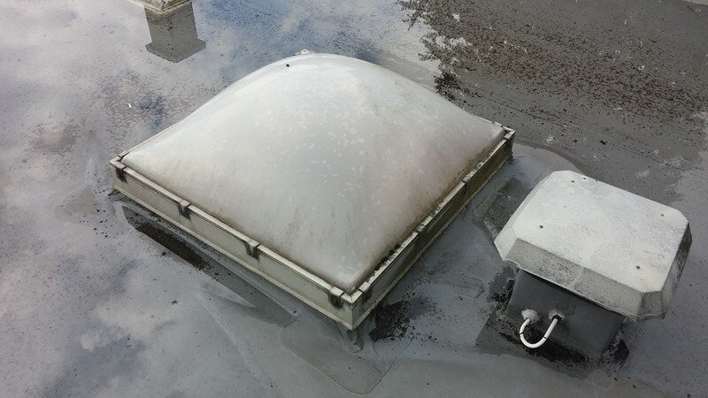 Nærbillede af kuppelformet ovenlys og en mindre ventilationshætte på et fladt tagpaptag med stående vand.