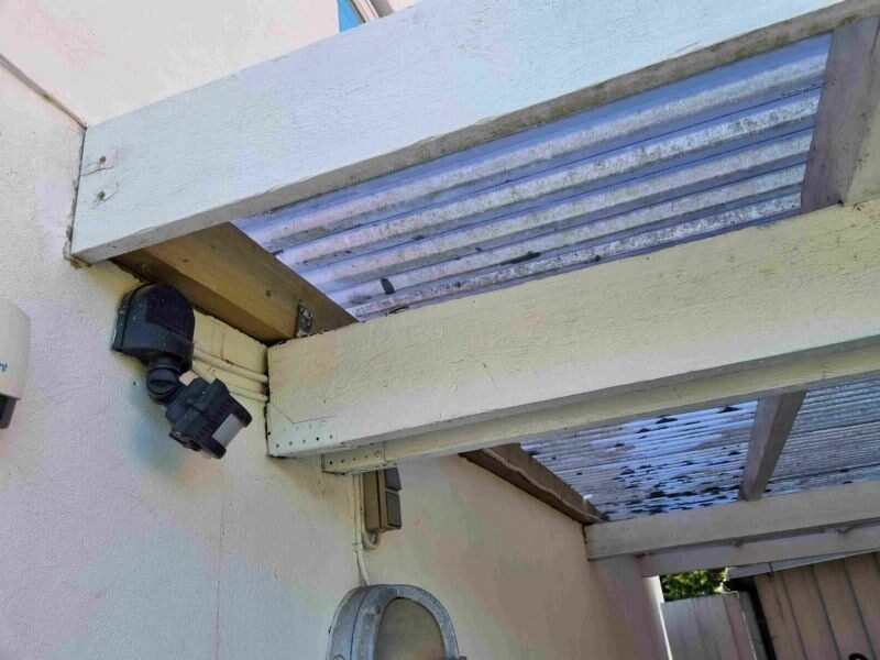 Nærbillede af snavset, gennemsigtig bølgeplade-overdækning på træbjælker ved en facade med udendørsbelysning.