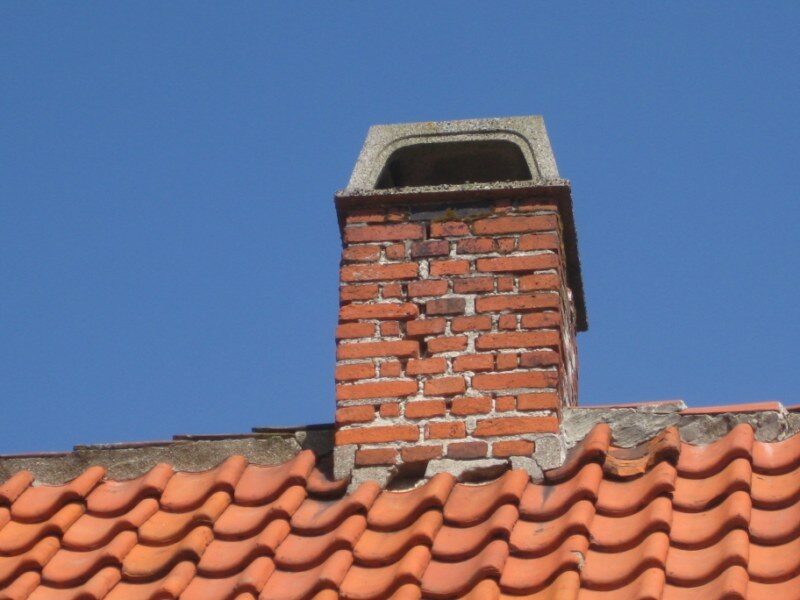 Nærbillede af en muret skorsten med betondæksel på et rødt tegltag mod klar blå himmel.