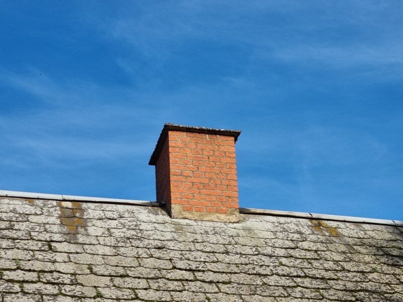 Nærbillede af en rød murstensskorsten på en tagryg med skifer- eller eternittag i blå himmel.