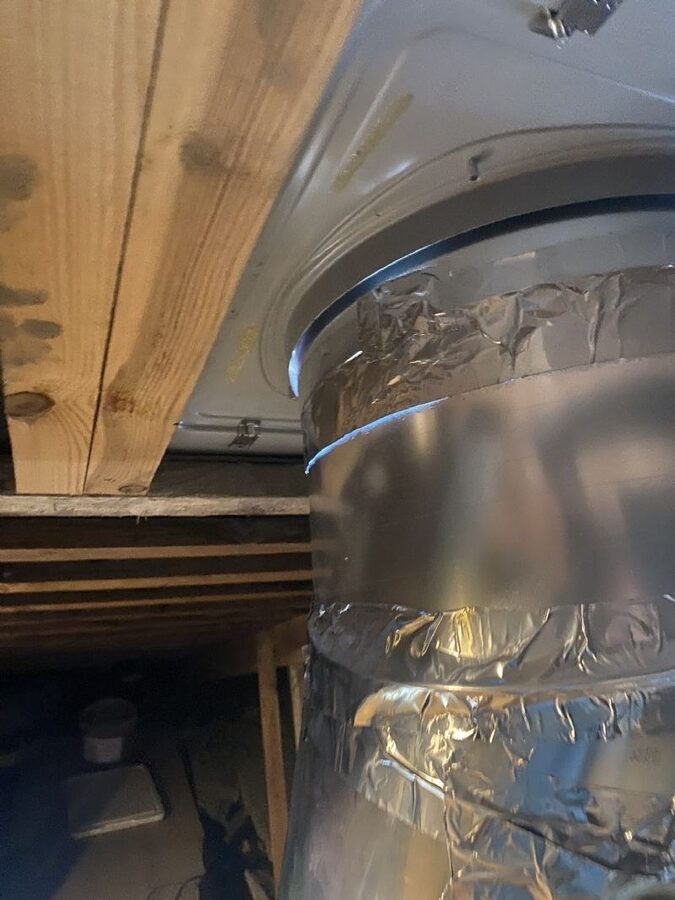 Nærbillede af en foliebeklædt ventilationskanal ført gennem en trækonstruktion i et tagrum eller kælderrum.
