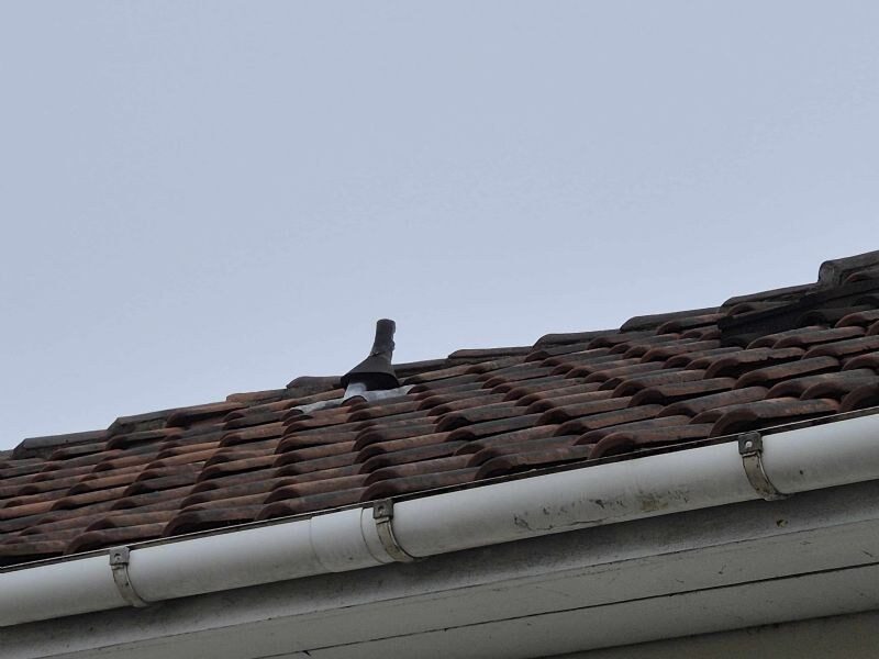 Nærbillede af tegltag med ventilationshætte og hvid tagrende, fotograferet nedefra mod himlen.