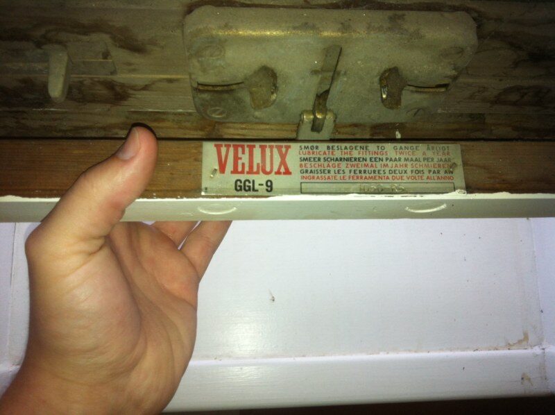 Nærbillede af et gammelt Velux ovenlysvindue GGL-9 med tydelige beslag og skiltetekst om smøring, holdt af en hånd.
