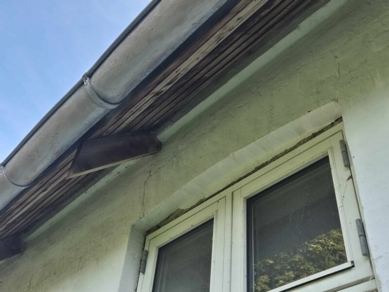 Nærbillede af udhæng med træbeklædning, zinktagrende og nedløb over et vindue på en pudset facade.
