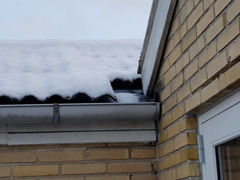 Snedækket tagkant med bølgeeternit, tagrende og gavl ses i nærbillede på gult murstenshus.