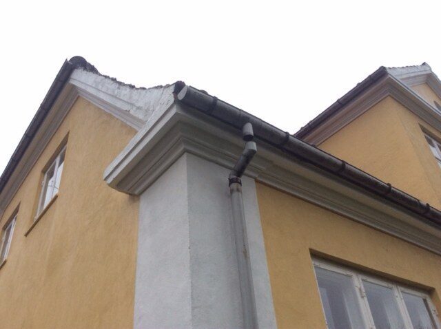 Nærbillede af gesims, tagrende og nedløb på gult pudset hus med skadet tagpapkant på taghjørnet.