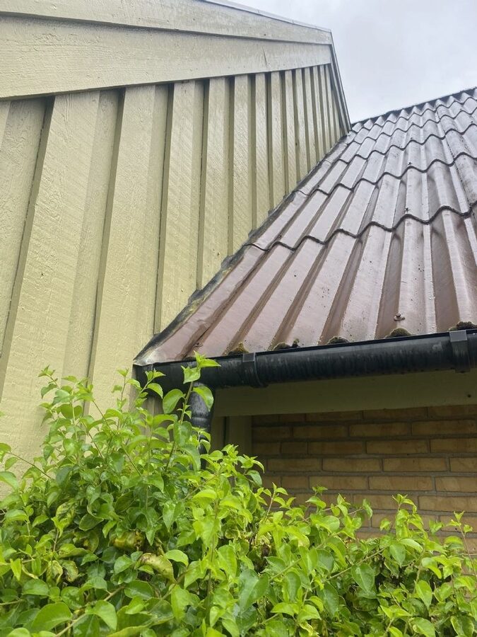 Nærbillede af tagfod med betontagsten, zinkinddækning og tagrende med let mosbegroning ved gavl beklædt med træbrædder.